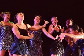 6 dancers in sparkly black dresses standing and gesturing next to each other.